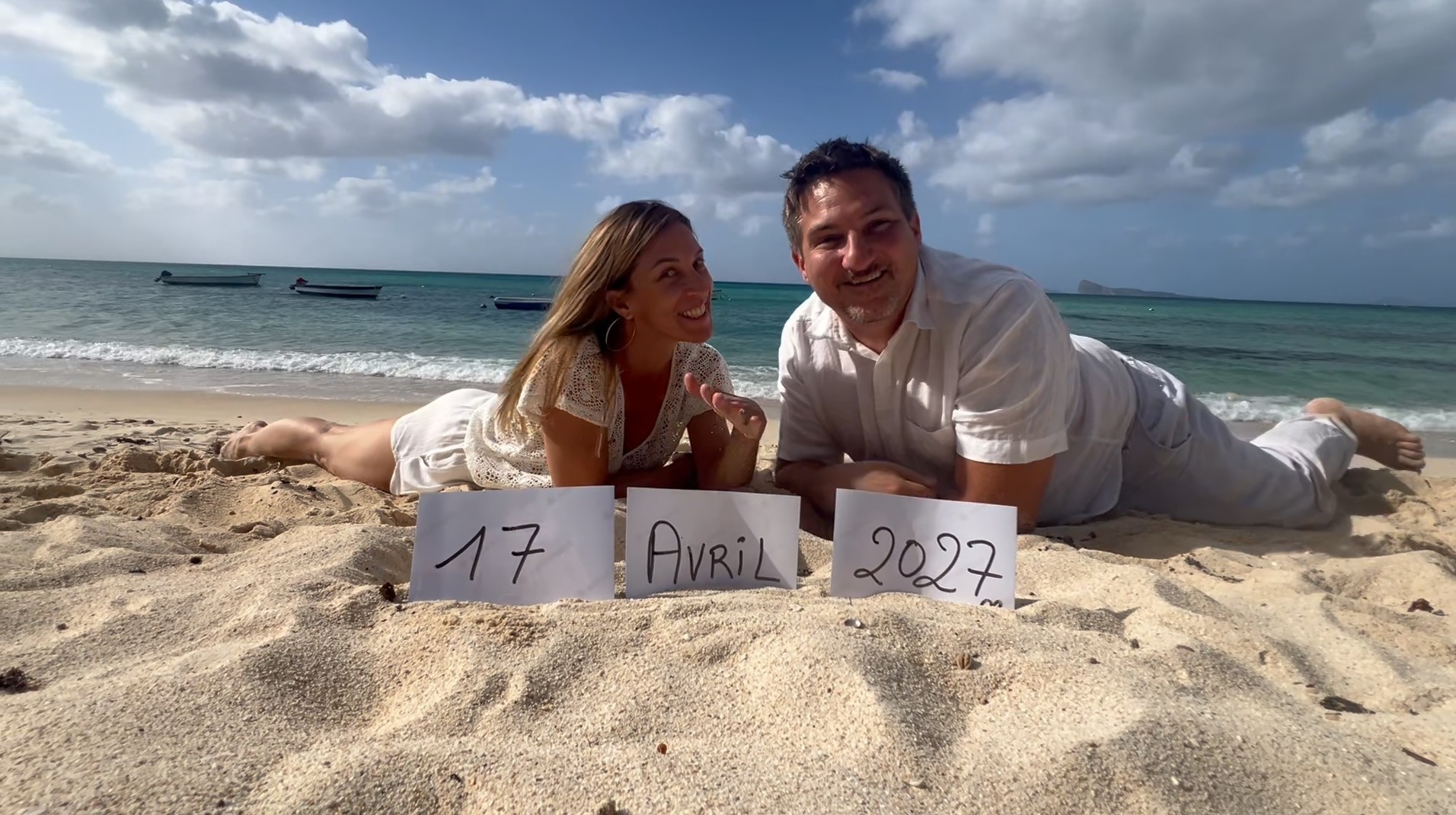 Florent et Isabelle allongés sur une plage de l'île Maurice, tenant des pancartes « 17 Avril 2027 », lagon turquoise en arrière-plan